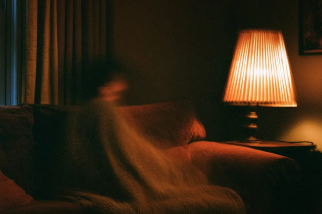 An empty sofa under a warm lamp, a blanket over the arm.