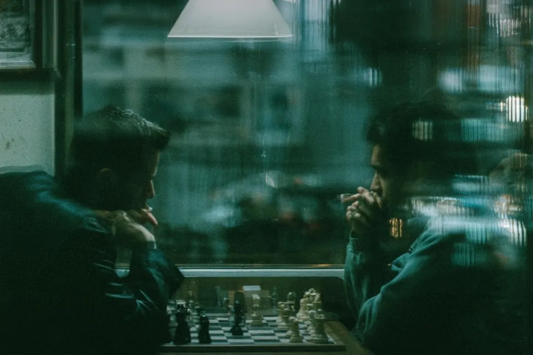 Two players over a chessboard, seen through a lit window.
