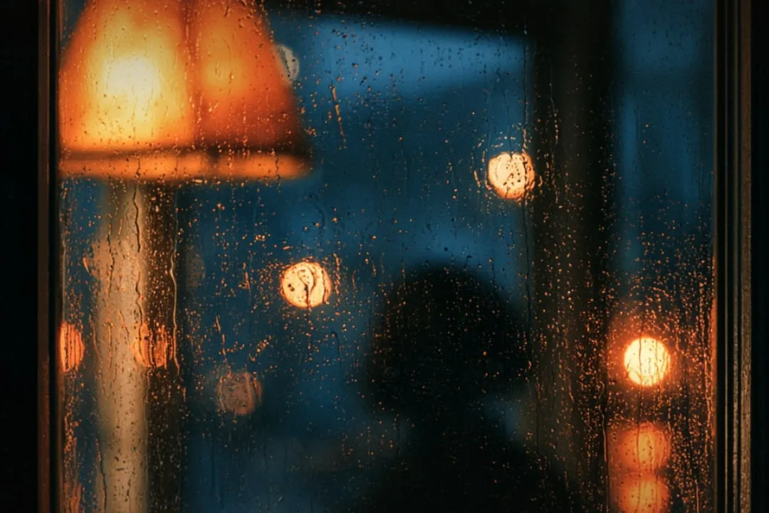 Rain running down a window, warm street lights blurred behind it.