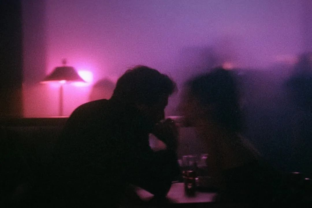 Two people leaning towards each other in a dim room, a pink lamp behind them.